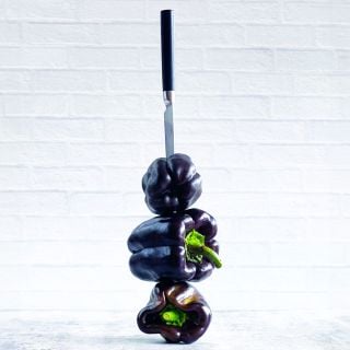 Three stacked Purple Beauty Peppers are held in place by a knife on a light background.