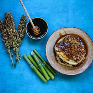 Seed heads and canes of Honey Drip Sorghum with sorghum syrup and pancakes on a bright blue background.