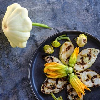 Grilled White Scallop Summer Squash with squash blooms as garnish on a metal background.