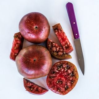 Cut and whole Black Krim Tomatoes arranged with a purple handled knife on white background.