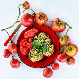Baked Mortgage Lifter Tomatoes on bright red plate surrounded with tomatoes one a white background.