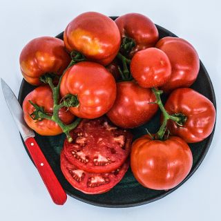 A pile of cut and whole Peron Tomatoes on dark ceramic plate with a red handled knife.