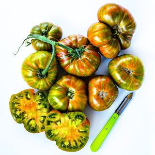 Seven whole Berkeley Tie-Dye Green Tomatoes arranged with a sliced tomato and green handled peeler.