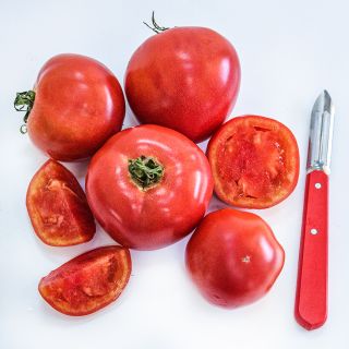 Whole, halved and quartered Ozark Pink Tomatoes with a red handled peeler on white background.