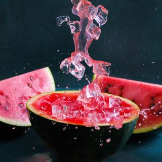 Iced watermelon juice splashing on top of a cut Sugar Baby Watermelon on a dark bluebackground.