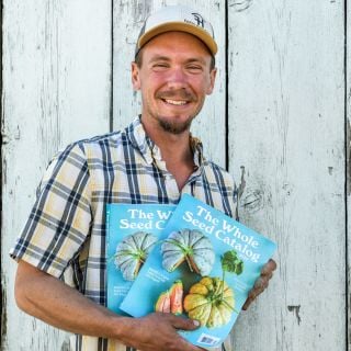 The Baker Creek farm manager smiles while holding a couple of 2025 Whole Seed Catalogs.